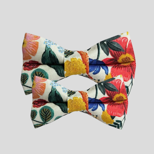 A colorful dog bow tie featuring the Rifle Paper Co. floral print with vibrant red, yellow, and pink flowers, green leaves, and blue accents on a white background. The bow tie has two elastic loops on the back for attaching to a collar, displayed against a plain light background.