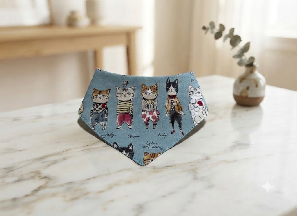 A triangular bandana-style pet bib with a light blue background and a printed pattern of dressed-up cats, displayed on a white marble tabletop in a softly lit indoor setting.