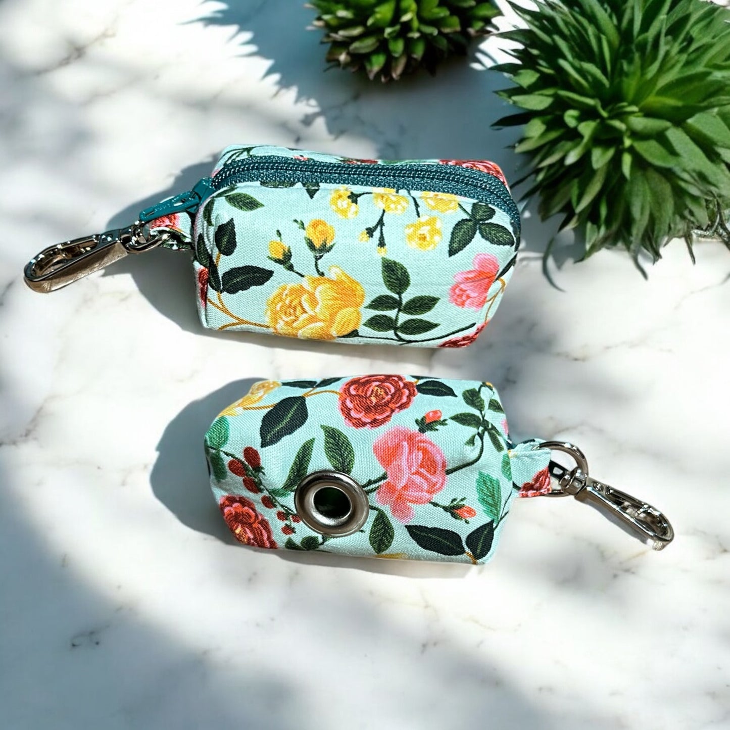Two small fabric pouches designed as dog poop bag holders, placed on a wooden surface. The pouch on the left has a blue zipper and features a colorful floral pattern with pink, yellow, and green flowers on a mint background, with a metal keychain clip. The pouch on the right has a metal eyelet and a similar floral design with pink, blue, and green flowers, also with a keychain clip. Both are likely handmade and suitable for carrying dog waste bags front and back.