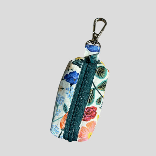A small, rectangular fabric pouch with a colorful floral pattern featuring blue, pink, and orange flowers, green leaves, and teal accents. The pouch has a green zipper and a metal clip attached to a short fabric loop at the top.