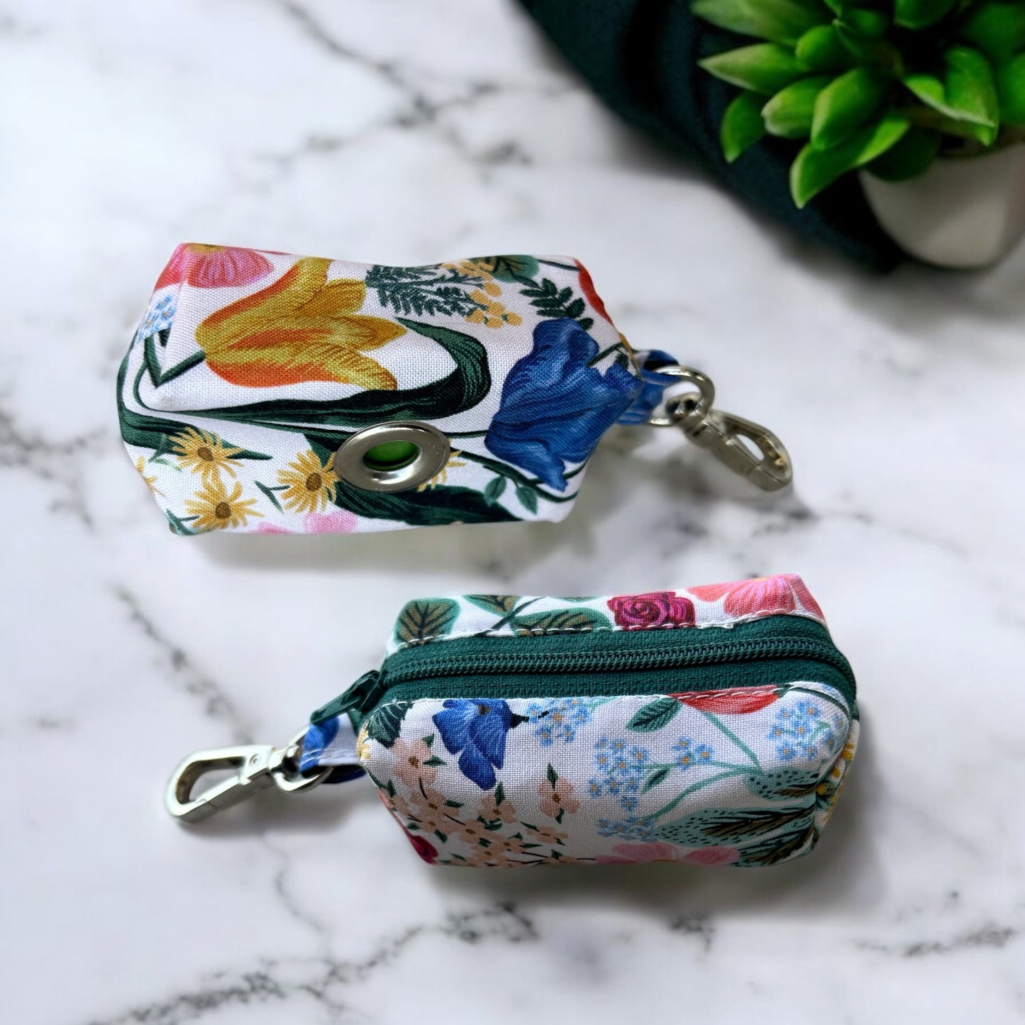 A small, rectangular fabric pouch with a colorful floral pattern featuring blue, pink, and orange flowers, green leaves, and teal accents. The pouch has a green zipper and a metal clip attached to a short fabric loop at the top.(back and side view