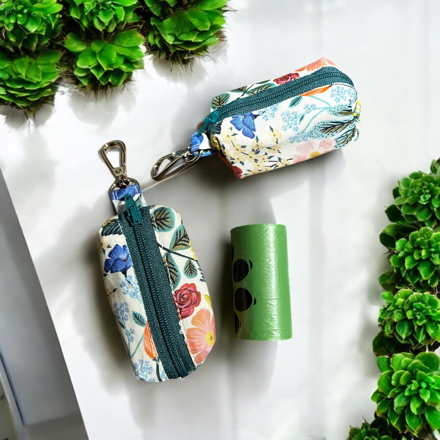 A small, rectangular fabric pouch with a colorful floral pattern featuring blue, pink, and orange flowers, green leaves, and teal accents. The pouch has a green zipper and a metal clip attached to a short fabric loop at the top side and front view with a roll of poop bags)