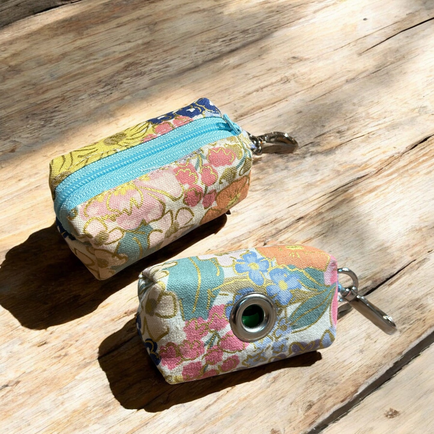 Two small fabric pouches designed as dog poop bag holders, placed on a wooden surface. The pouch on the left has a blue zipper and features a colorful floral pattern with pink, yellow, and green flowers on a cream background, with a metal keychain clip. The pouch on the right has a metal eyelet and a similar floral design with pink, blue, and green flowers, also with a keychain clip. Both are likely handmade and suitable for carrying dog waste bags.