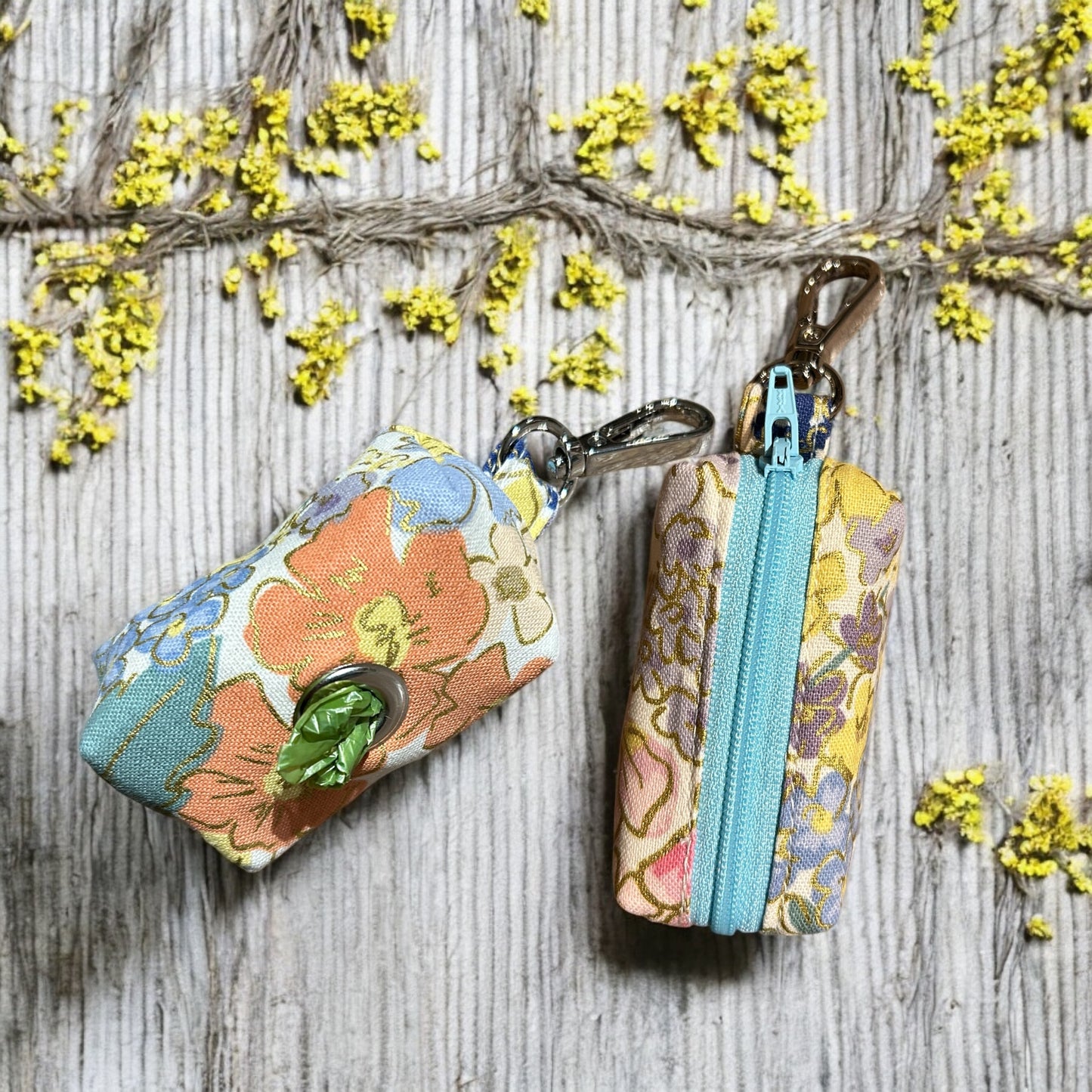 Two small fabric pouches designed as dog poop bag holders, placed on a wooden surface. The pouch on the left has a blue zipper and features a colorful floral pattern with pink, yellow, and green flowers on a cream background, with a metal keychain clip. The pouch on the right has a metal eyelet and a similar floral design with pink, blue, and green flowers, also with a keychain clip. Both are likely handmade and suitable for carrying dog waste bags.