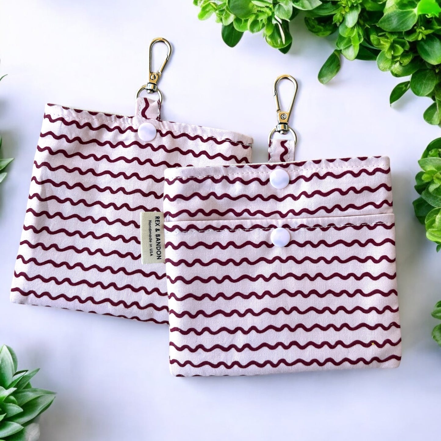 A small fabric pouch with a floral design featuring pink, red, and yellow roses, green leaves, and red berries on a light green background. The pouch has a snap closure, a metal keychain clip attached to a fabric loop, and a "Handmade in USA" label (front view)