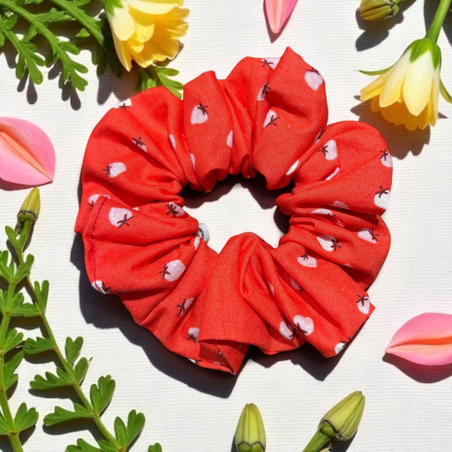 Red scrunchie with white heart pattern on a light background with flowers and leaves.