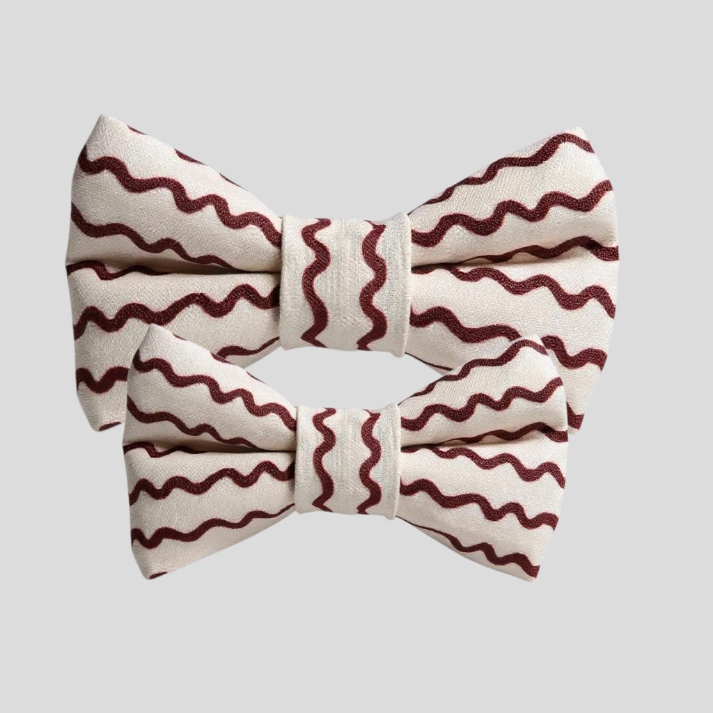 A stylish dog bow tie with a ripple pattern in deep maroon on a creamy white background. The bow tie features two elastic loops on the back for attaching to a collar, displayed against a plain light background rifle paper co.