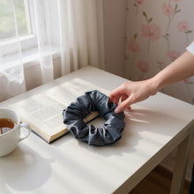 Load image into Gallery viewer, Hand reaching for a eggplant scrunchie on a table with an open book and cup of tea.