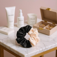 Load image into Gallery viewer, Black and beige scrunchies on a marble surface with skincare products and jewelry box in the background.