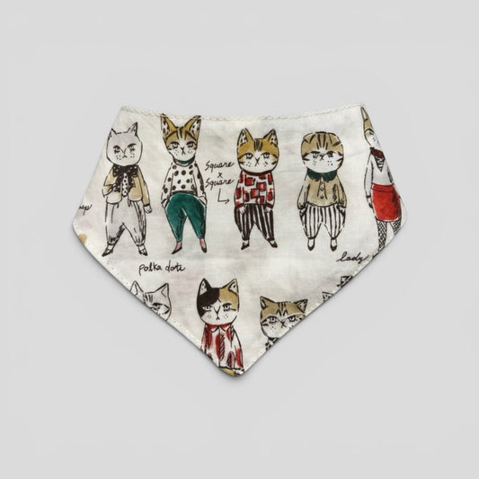 A triangular bandana bib with an off-white background printed with a whimsical pattern of standing cats in different outfits and poses.