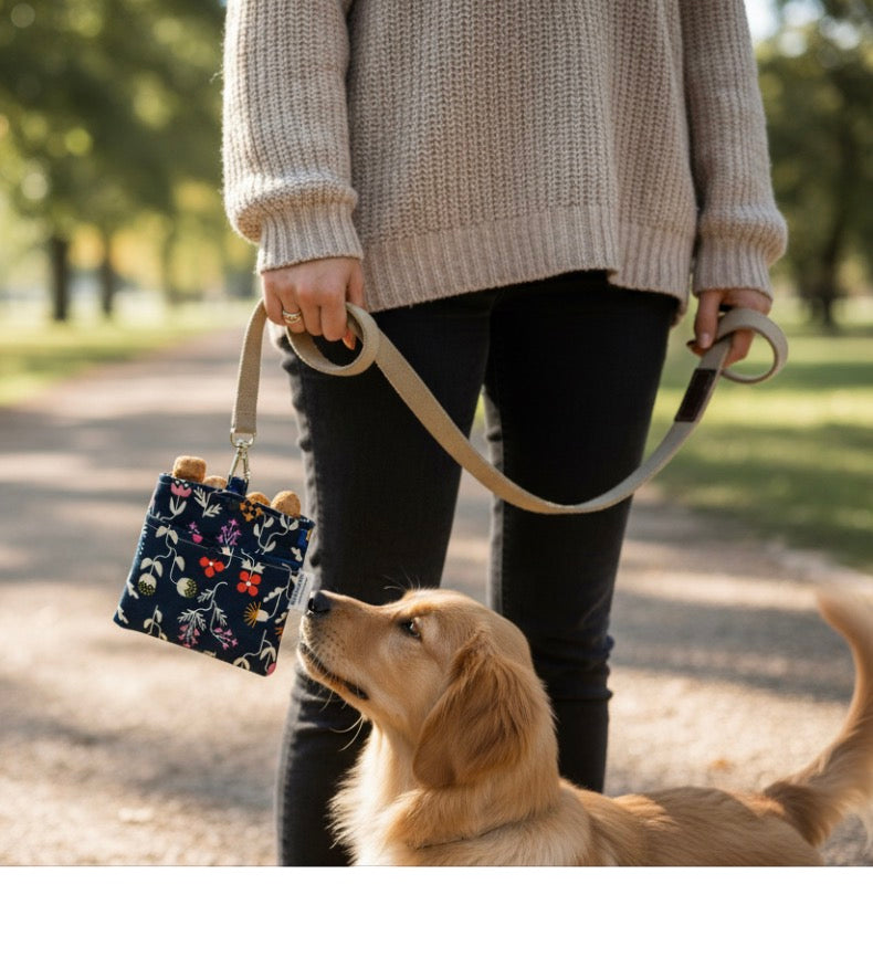 A person in a beige knit sweater and black pants holds a small floral dog leash bag while walking a golden retriever on a path in a park.