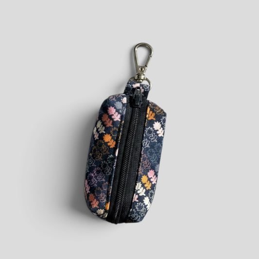 A small zippered pouch with a floral print in navy, pink, orange, and white, featuring a black zipper and a silver metal carabiner clip, shown on a light gray background.