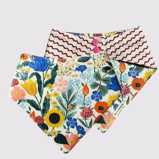 A reversible dog bandana with one side featuring the Rifle Paper Co. GardenWhimsy print with colorful flowers and greenery on a white background, and the reverse side displaying the GardenRipple print with maroon wave patterns on a creamy background. The bandana has snap closures, shown against a plain light background.