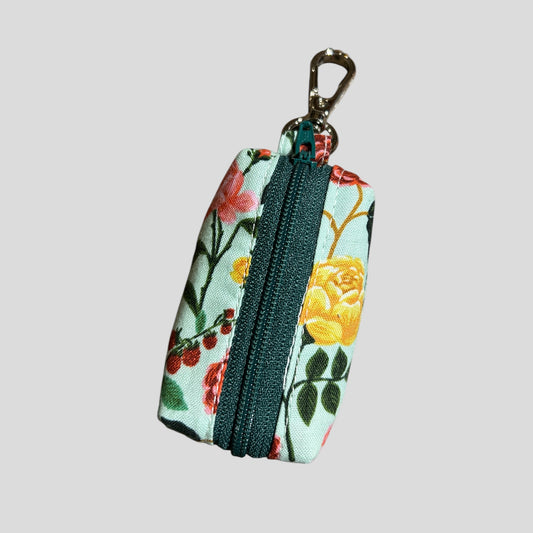 A small fabric dog poop bag holder by Rifle Paper Co. in the ShimmerGrande design, featuring a vibrant floral pattern with pink, yellow, and green flowers on a cream background with gold accents. The pouch has a blue zipper and a metal keychain clip, ideal for carrying waste bags while walking a pet. front view