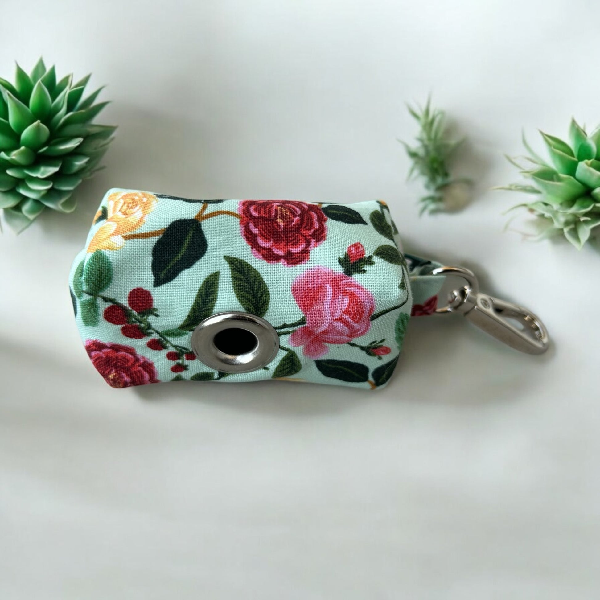 A small fabric dog poop bag holder by Rifle Paper Co. in the ShimmerGrande design, featuring a vibrant floral pattern with pink, yellow, and green flowers on a cream background with gold accents. The pouch has a blue zipper and a metal keychain clip, ideal for carrying waste bags while walking a pet. Back view