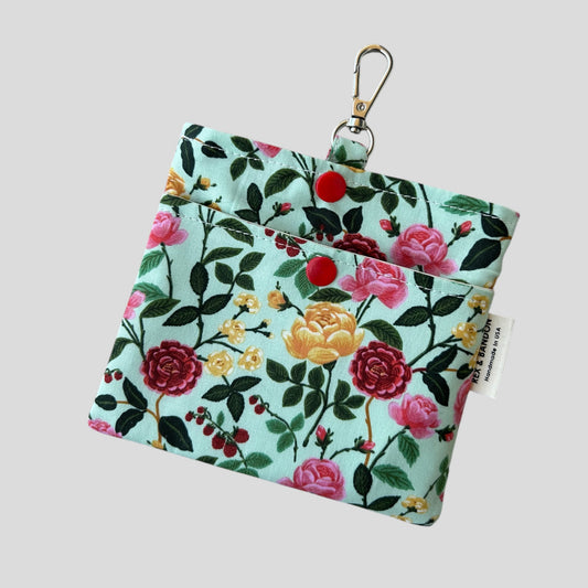 A small fabric pouch with a floral design featuring pink, red, and yellow roses, green leaves, and red berries on a light green background. The pouch has a snap closure, a metal keychain clip attached to a fabric loop, and a "Handmade in USA" label.