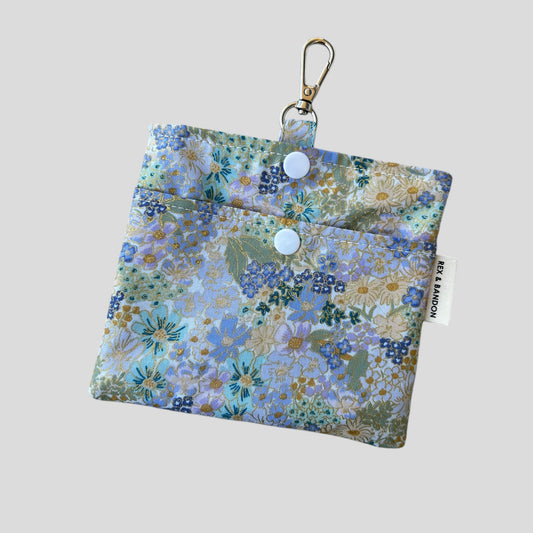 A floral printed dog treat bag with a mix of pastel colors and metallic shimmer, featuring a front pocket, snap buttons, and an alloy metal clip.