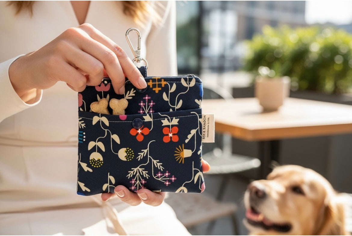 A person holds a small floral pet carrier wallet with a metal clip and a small dog tag, with a golden retriever dog looking on in a sunny outdoor setting.