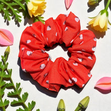 Load image into Gallery viewer, Red scrunchie with white heart pattern on a light background with flowers and leaves.