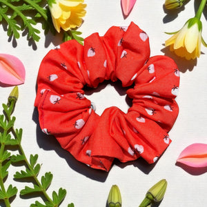 Red scrunchie with white heart pattern on a light background with flowers and leaves.