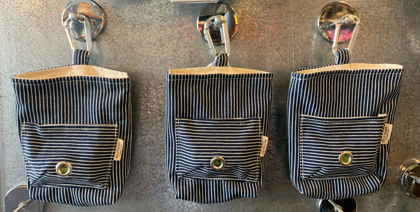 A striped cotton dog walker pouch with a front pocket and a carabiner clip, hanging 