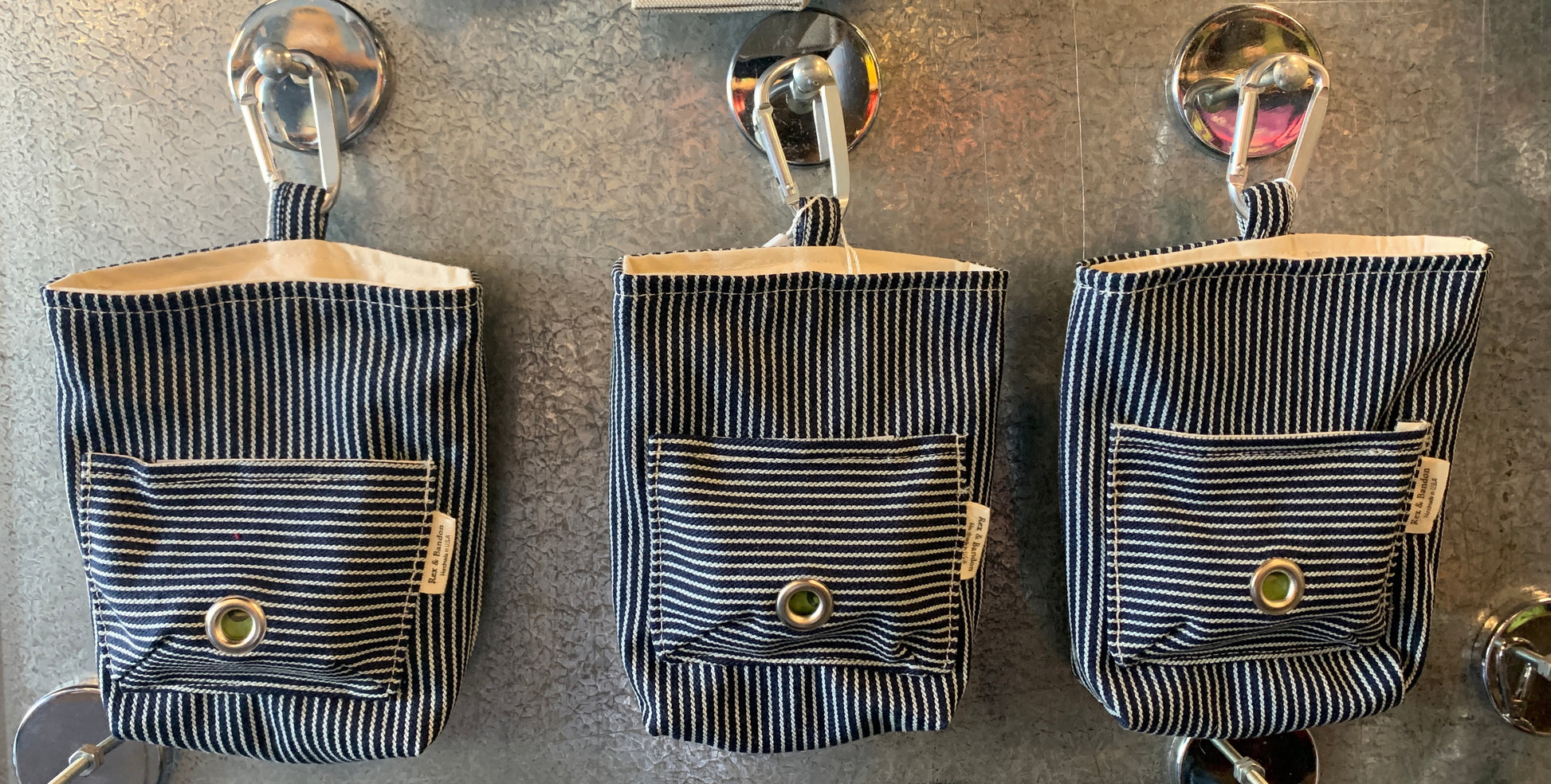 A striped cotton dog walker pouch with a front pocket and a carabiner clip, hanging 